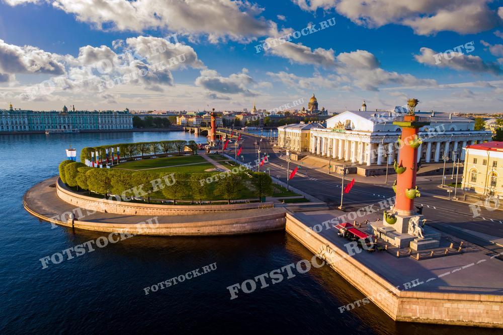 На фото изображена панорама Санкт-Петербурга с высоты птичьего полета. На переднем плане видна Стрелка Васильевского острова с Ростральными колоннами и зданием Биржи. На заднем плане расположены Зимний дворец и Эрмитаж, а также купол Исаакиевского собора.